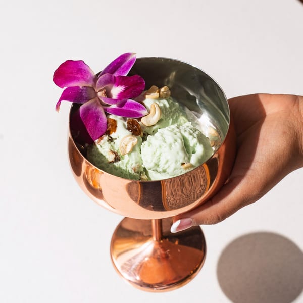 Hand holding a copper goblet of green ice cream topped with cashews, raisins, and an orchid.