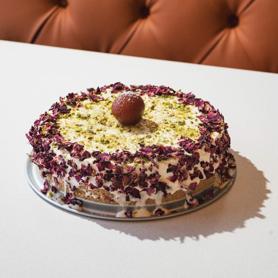 Indian dessert cake with white frosting, rose petals, pistachios, and a gulab jamun on top.