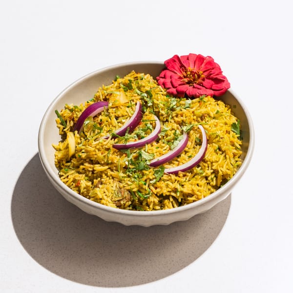 A bowl of yellow Indian rice, garnished with red onion slices, cilantro, and a red flower.