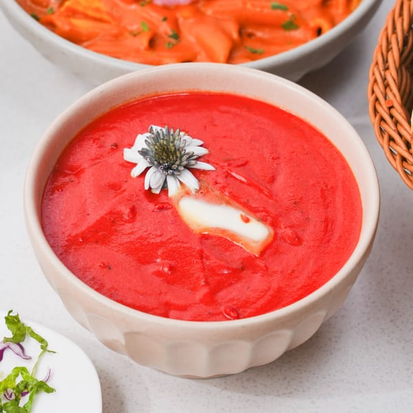 A bowl of vibrant red Indian tomato soup, garnished with a white flower and a pat of butter.