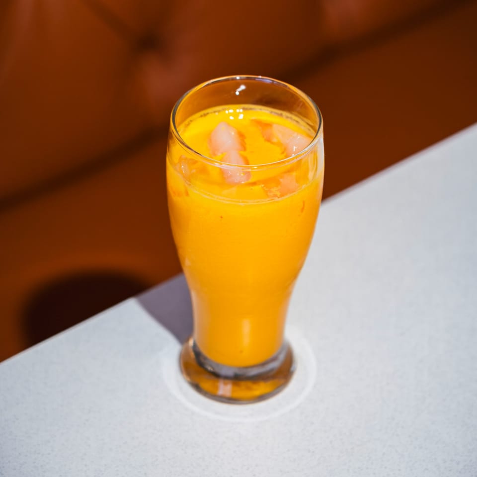 A vibrant orange mango lassi, an Indian drink, in a tall glass with fruit chunks on a white table.