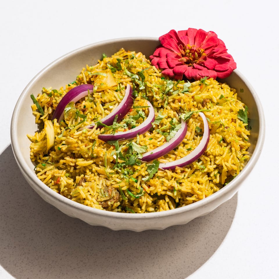 A bowl of yellow Indian rice, garnished with red onion slices, cilantro, and a red flower.
