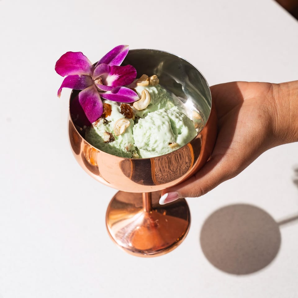 Hand holding a copper goblet of green kulfi, topped with cashews, raisins, and an orchid.