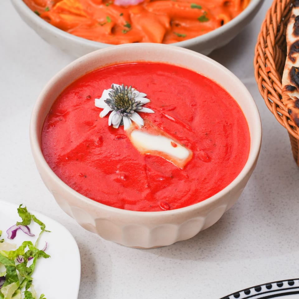 A bowl of vibrant red Indian curry, garnished with a white flower and a dollop of butter.