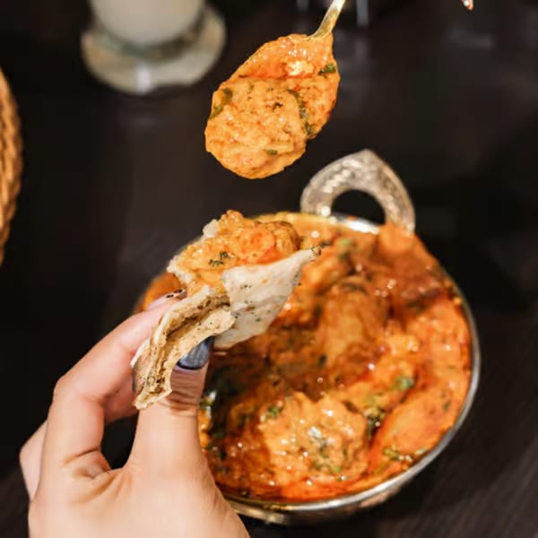 A hand holding naan bread scoops Indian curry from a silver bowl, with a spoonful of curry above.