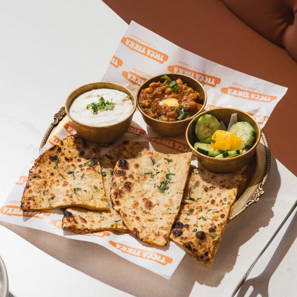 A golden tray holds naan bread, chickpea curry, raita, and cucumber salad from Ticca Tikka.