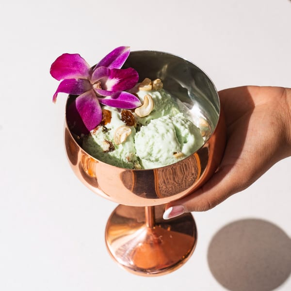 Hand holding a copper goblet filled with green pistachio kulfi, topped with cashews, raisins, and an orchid.