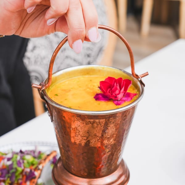 A hand holds a copper bucket of yellow Indian curry with a red flower garnish.