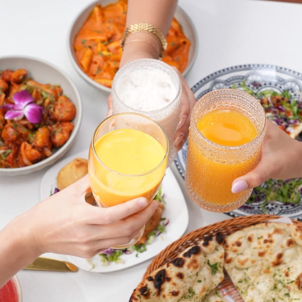 Four hands toasting with mango lassi and other drinks, surrounded by Indian dishes like naan and curry.
